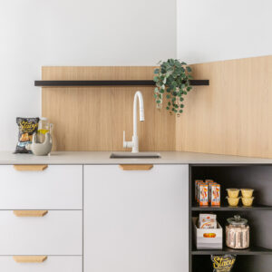 A modern kitchenette and snack station featuring sleek white flat-panel cabinets with light wood edge pulls and a smooth light-colored countertop. A modern white gooseneck faucet and under-mount sink sit beneath a warm light wood paneled backsplash and a minimalist black floating shelf styled with a trailing green plant. To the right, dark open shelving displays neatly organized snacks and drinks against crisp white walls.