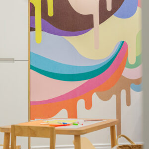 A vibrant children's playroom featuring a large, colorful abstract mural with a fun, dripping paint design in bright pinks, blues, yellows, and browns. In the foreground, a small light wood kids' art table and chair set is laid out with paper and markers, alongside a woven storage basket with white pom-poms.