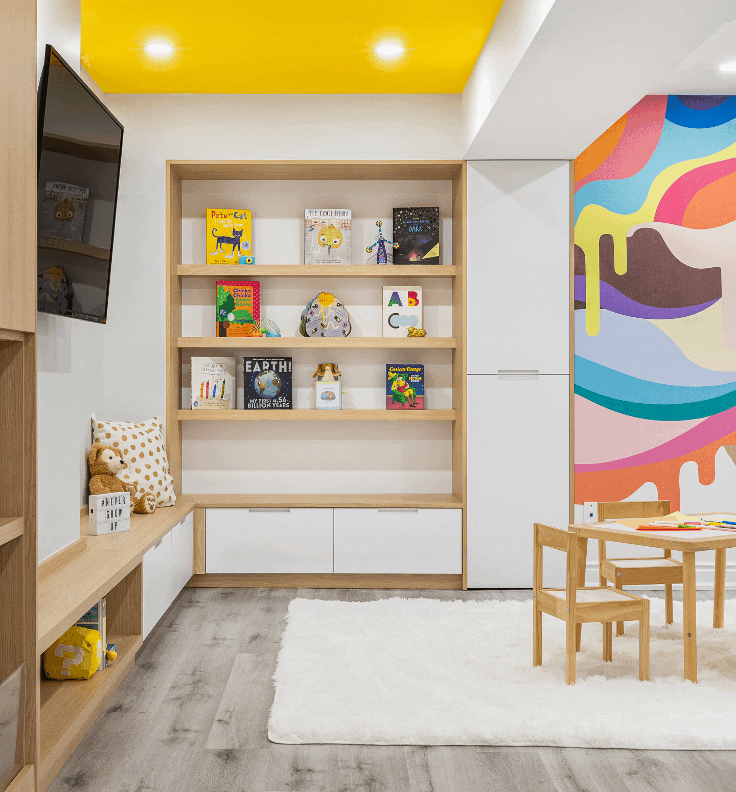 A bright and energetic kids' playroom featuring custom light wood built-in bookshelves displaying front-facing children's books. Below the shelves is an L-shaped bench with white storage drawers, styled with a plush toy and a patterned pillow. The space is highlighted by a vibrant yellow painted ceiling and a colorful abstract drip mural on the right wall. A small wooden children's table and chair set sits near a black three-tier rolling art cart, all resting on a soft white area rug over light wood-look flooring.