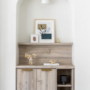 A modern white arched wall niche featuring custom light wood built-in cabinetry. The lower unit includes two cabinet doors with sleek gold edge pulls and open side shelving displaying candles and a small black vessel. The wood counter and matching backsplash panel are styled with a vase of dried florals and a book, while framed abstract art rests on a floating wood shelf above. A contemporary white and brass wall sconce illuminates the top of the alcove.
