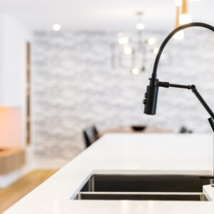 A close-up view of a modern, matte black articulating pull-down kitchen faucet and matching soap dispenser mounted on a bright white quartz island countertop. Below it is a stainless steel double-bowl under-mount sink. The softly blurred background reveals an open-concept dining and living space featuring a modern sputnik chandelier, a textured accent wall, and a glimpse of a linear fireplace.