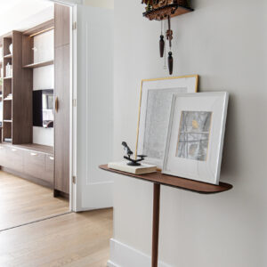A bright hallway featuring a unique, minimalist single-leg wooden console table resting against a light neutral wall. The narrow shelf is elegantly styled with two layered framed art prints, a book, and a small black bird sculpture. A traditional dark wood cuckoo clock hangs on the wall above. To the left, open white doors reveal a glimpse into a living room featuring floor-to-ceiling dark wood built-in cabinetry and light hardwood floors.