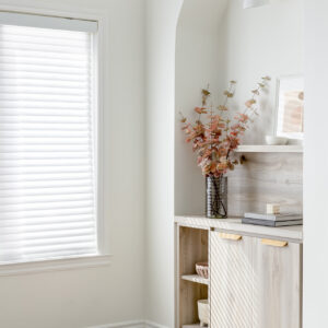 A bright corner featuring a white arched wall niche with custom light wood built-in cabinetry. The lower unit includes closed doors with modern gold edge pulls and open side shelving, topped with a matching wood backsplash. The counter is elegantly styled with a tall glass vase holding faux branches, stacked books, and a bubble candle. A modern white and brass wall sconce illuminates the upper alcove. Sunlight streams through an adjacent window with white blinds, casting sharp linear shadows across the cabinets and light wood floor.
