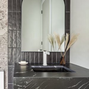A stylish modern bathroom vanity featuring a thick black marble-look countertop with striking white veining and a seamless integrated sink. A sleek white modern faucet sits in front of a backsplash made of vertically stacked, textured black tiles. An elegant arched mirror with a thin black frame hangs above. The counter is accessorized with a small amber glass vase holding dried decorative grasses, with a glimpse of patterned terrazzo wall tile to the left.