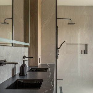A sleek modern bathroom featuring a double vanity with a dramatic black stone countertop with white veining and integrated square sinks. Two matte black wall-mounted faucets sit beneath a large rectangular LED-illuminated mirror. To the right, a walk-in shower with large-format light grey wall tiles features a black-framed glass enclosure, matte black shower fixtures, and a recessed product niche.