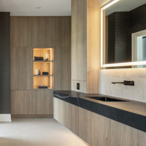A luxurious modern bathroom featuring a long floating light wood vanity topped with a thick black stone countertop with white veining and an integrated sink. A sleek matte black wall-mounted faucet is installed beneath a large, rectangular LED backlit mirror. In the background, seamless floor-to-ceiling matching wood cabinetry includes a beautifully illuminated recessed display niche styled with dark folded towels and candles.