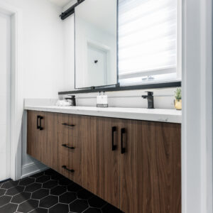 A bright, modern bathroom featuring a long floating dark wood double vanity with a white marble-look quartz countertop and matte black cabinet pulls and faucets. The floor is finished with striking large matte black hexagonal tiles with contrasting white grout. Above the sinks, a large rectangular mirror slides on a black industrial barn-door track, partially covering a window fitted with white horizontal blinds.