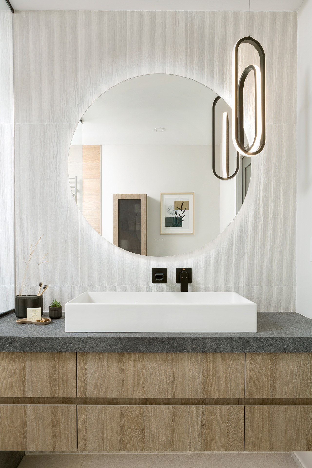 A modern bathroom featuring a floating light wood vanity with a dark textured stone countertop and a white rectangular vessel sink. A minimalist matte black wall-mounted faucet sits beneath a large round backlit mirror on a vertically textured white wall, accented by a contemporary black oval pendant light.