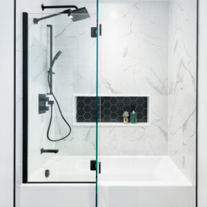 A modern, high-contrast bathroom featuring a white bathtub and shower combination with a partial frameless glass screen and black hardware. The shower surround is finished in large-format white marble-look tiles, complete with a built-in horizontal niche backed by black hexagonal tiles. Sleek matte black plumbing fixtures, including a rain shower head and handheld wand, stand out against the white walls, while the floor is laid with striking matte black hexagonal tiles with white grout.