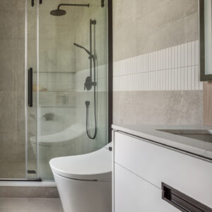 A contemporary bathroom featuring a sleek white floating vanity with integrated black hardware and a modern white skirted toilet. To the left, a walk-in shower is enclosed by a sliding glass door with a matte black track and matching black shower fixtures. The walls and floor are finished in large-format light grey stone-look tiles, beautifully accented by a horizontal band of white vertical kitkat tiles.
