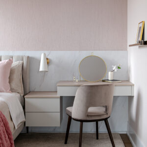 A chic modern bedroom corner featuring a custom white floating nightstand that seamlessly steps up into a built-in vanity desk, both accented with light wood tops. The vanity area is elegantly styled with a round gold mirror and paired with a grey velvet chair with dark wood legs. The backdrop showcases a striking two-toned wall with a white marble-look lower panel and a textured soft pink upper section, illuminated by a modern brass and white cone wall sconce.