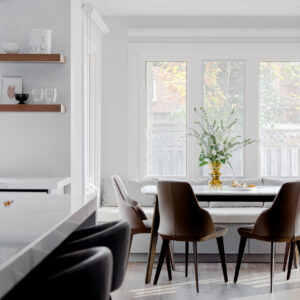 A bright, modern kitchen breakfast nook featuring a built-in banquette window seat with light grey cushions nestled beneath a large three-pane window. A sleek rectangular dining table is surrounded by four contemporary curved brown leather dining chairs. To the left, a glimpse of the kitchen reveals a white quartz island with dark grey bar stools and a wall corner elegantly styled with two dark wood floating shelves.