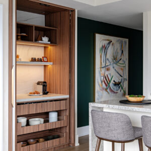 A modern home interior featuring a custom floor-to-ceiling built-in coffee bar with fully retracted pocket doors. The warm wood-lined interior showcases integrated LED lighting, open shelving, and a sleek stone backsplash and countertop holding a coffee maker and pastries. Below the counter, three open pull-out wood drawers neatly store various bowls and dishes. To the right, a stone-topped kitchen island with grey upholstered wooden bar stools sits in front of a dark green accent wall displaying a large abstract painting.