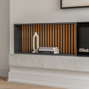 A close-up detail of a modern living room featuring a dark grey built-in display niche with a warm vertical wood slat backing. The shelf is minimally styled with a white ceramic loop sculpture and a small stack of books. Below, a thick floating white marble hearth extends along the wall over light oak chevron flooring and the edge of a fluffy white area rug. A partial view of a black linear fireplace is visible on the right.