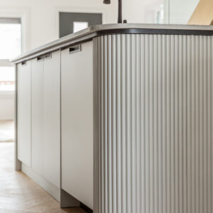 A close-up detail shot of a modern kitchen island featuring a distinctive rounded end covered in light grey vertical fluted paneling. The side of the island showcases sleek, flat-panel cabinetry with minimalist black edge pulls and a light stone countertop. In the softly blurred background, a matte black gooseneck faucet and light oak chevron wood flooring complete the contemporary design.
