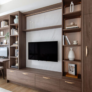 A modern custom built-in entertainment center crafted from warm dark wood. The floor-to-ceiling unit features a central flat-screen TV mounted against a sleek white marble-look backdrop, flanked by illuminated open shelving displaying books and minimalist white ceramics. Below the TV, seamless drawers are accented with contemporary gold hardware. A built-in desk area seamlessly integrates into the left side of the unit, all set above light hardwood floors and a textured white area rug.