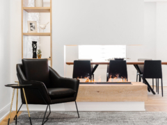 A modern living area featuring a sleek black leather accent chair and a small black and gold side table positioned near a striking double-sided linear fireplace. The fireplace sits on a chunky light wood base beneath a white floating partition wall, acting as a stylish room divider that offers a clear view into the adjacent dining room with black chairs and a long table. To the left, a floor-to-ceiling light wood open shelving unit is elegantly styled with minimal decor.
