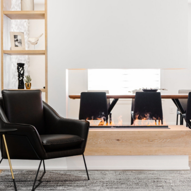 A modern living area featuring a sleek black leather accent chair and a small black and gold side table positioned near a striking double-sided linear fireplace. The fireplace sits on a chunky light wood base beneath a white floating partition wall, acting as a stylish room divider that offers a clear view into the adjacent dining room with black chairs and a long table. To the left, a floor-to-ceiling light wood open shelving unit is elegantly styled with minimal decor.
