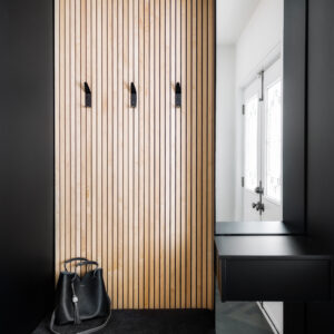 A modern, custom entryway nook featuring a built-in matte black bench with a dark upholstered seat cushion, set against a striking light wood vertical slat accent wall with three minimalist black coat hooks. A black leather tote bag rests on the bench. To the right, a sleek black floating drawer and countertop sit beneath a tall mirror, which reflects a bright white front door with glass details and dark grey herringbone floor tiles.