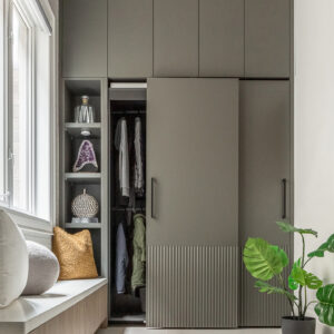A modern entryway featuring custom floor-to-ceiling cabinetry in a matte olive-grey finish. The sliding wardrobe doors showcase a stylish fluted texture on the lower half and sleek black pull handles, with one door partially open revealing hanging coats. To the left, a built-in light wood window bench with a white countertop and decorative pillows sits next to open vertical shelving displaying a crystal and vases. A potted monstera plant rests on the light neutral tile floor to the right.