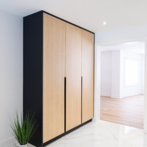 A bright, modern hallway featuring a large custom built-in wardrobe with a matte black frame and sleek light wood flat-panel doors accented by long black vertical pulls. The space is anchored by polished large-format white marble-look floor tiles. A small potted green grass plant sits to the left of the closet, while an open doorway to the right reveals an adjacent bright room with light wood flooring."