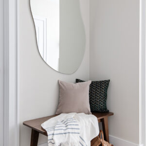 A cozy entryway corner featuring a sleek dark wood bench styled with a solid beige pillow, a dark green geometric patterned pillow, and a white throw blanket with blue stripes. Above the bench hangs a large, frameless organic-shaped mirror. A woven wicker basket sits on the light wood floor beneath the bench, all set against soft, light neutral walls with crisp white trim.