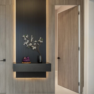 A contemporary hallway entryway featuring seamless floor-to-ceiling light wood paneling with an integrated flush-mount door. The focal point is a dramatic, matte black fluted wood accent panel, highlighted by warm LED backlighting. A chunky black floating console shelf extends from the panel, beautifully styled with a dark vase holding white floral branches and a small purple decorative object.