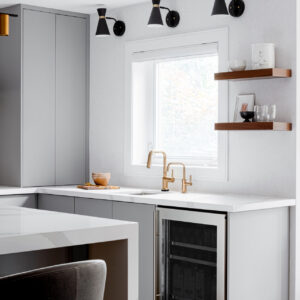 A bright, modern kitchen area featuring light grey flat-panel base cabinets and a white marble-look quartz countertop equipped with an elegant brass faucet and a built-in stainless steel beverage fridge. Three black mid-century style cone wall sconces are mounted above the sink window. To the right, two dark wood floating shelves display minimalist ceramics and glassware. A partial view of a quartz waterfall island and a dark grey velvet bar stool is visible in the foreground over light wood flooring.