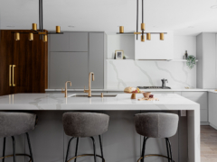 A sleek, contemporary kitchen featuring a large white quartz island with a bold grey-veined waterfall edge, dark grey base cabinets, and elegant brass faucets. Three grey velvet bar stools sit at the counter beneath two modern black and gold linear chandeliers. The background showcases minimalist light grey flat-panel cabinetry, a matching continuous quartz backsplash with a floating shelf, and a contrasting dark wood-paneled built-in refrigerator with long gold handles.