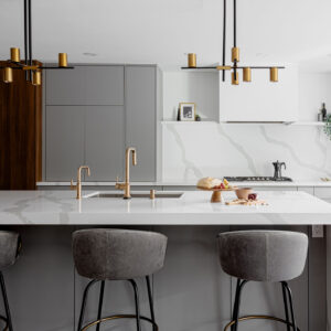 A sleek, contemporary kitchen featuring a large white quartz island with a bold grey-veined waterfall edge, dark grey base cabinets, and elegant brass faucets. Three grey velvet bar stools sit at the counter beneath two modern black and gold linear chandeliers. The background showcases minimalist light grey flat-panel cabinetry, a matching continuous quartz backsplash with a floating shelf, and a contrasting dark wood-paneled built-in refrigerator with long gold handles.