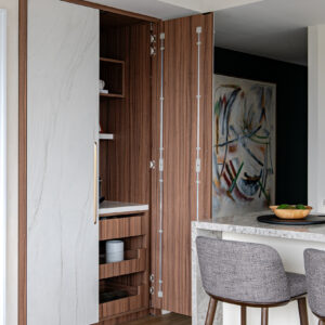 A modern home interior showcasing a custom floor-to-ceiling hidden bar cabinet. The unit features space-saving pocket doors with a white marble-look finish and sleek gold hardware. One door is retracted, revealing a warm wood interior equipped with open shelving, a sleek countertop, and pull-out storage drawers. To the right, a stone-topped kitchen island with grey upholstered wooden bar stools sits in front of a dark green accent wall displaying a large abstract painting.