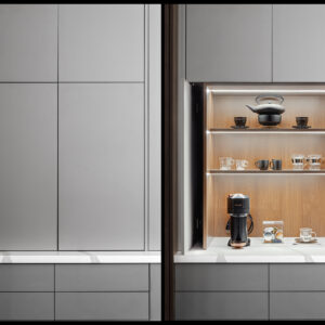A modern hidden kitchen coffee and tea bar revealed by opened pocket doors within sleek, grey flat-panel cabinetry. The illuminated appliance garage features a warm wood-paneled interior with two wood shelves glowing with integrated LED strip lighting. A black teapot set rests on the top shelf, various glass and black mugs sit on the middle shelf, and a modern black coffee maker with pod storage is ready for use on the white marble-look quartz countertop below.