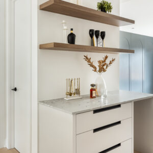 A stylish modern home bar nook featuring white drawer cabinetry with sleek black edge pulls and a speckled light grey stone countertop. Two thick wood floating shelves are mounted on the crisp white wall above, elegantly styled with black wine glasses, candles, and a small potted plant. The counter is accessorized with gold bar tools, a liquor bottle, and a white vase with dried foliage, all resting on light oak chevron wood flooring.
