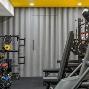 A modern home gym featuring a grey vertical fluted accent wall with a seamlessly integrated hidden door and a sleek black handle. A bold yellow painted ceiling brings energy to the space. The room is fully equipped with professional fitness gear, including a dumbbell rack with colorful weights, a Rogue power rack with a weight bench, and a Matrix cardio machine in the foreground, all resting on black rubber gym flooring.