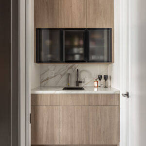 A stylish built-in wet bar tucked into a hallway alcove. The design features flat-panel light wood cabinetry on the top and bottom, separated by three striking black-framed smoked glass upper cabinets. A white marble-look quartz slab is used for both the countertop and continuous backsplash, complete with a small undermount sink, a sleek black faucet, and a silver water dispenser.
