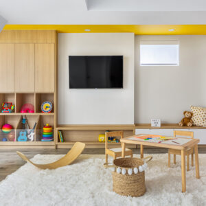 Project: CorduroyA bright, modern children's playroom featuring extensive custom light wood built-in cabinetry with open cubbies, shelves, and low bench storage. A flat-screen TV is mounted on the white wall beneath a striking vibrant yellow painted ceiling accent. The play area is centered on a large, plush white area rug and includes a small wooden kids' table and chairs, a curved wooden balance board, and a woven storage basket.