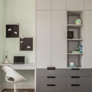 A modern kid's study area featuring a custom built-in unit with light grey upper cabinets, dark grey lower drawers with black hardware, and open shelving displaying books and plush toys. To the left, a minimalist white floating desk holds a laptop against a soft mint green wall, paired with a modern white rolling chair and two black textured pinboards.