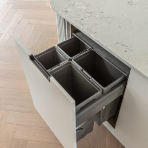 A top-down view of an open pull-out waste and recycling drawer integrated into a modern kitchen island. The sleek, light grey flat-panel cabinet features a minimalist black edge pull and houses a convenient four-bin sorting system. The island is topped with a light grey speckled stone counter with rounded corners, set beautifully against light oak chevron wood flooring.