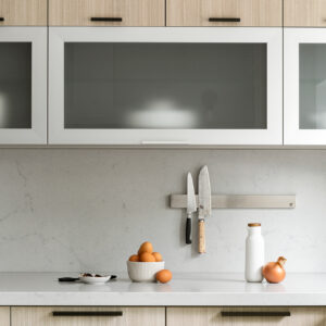 A close-up view of a modern kitchen featuring flat-panel light wood cabinetry accented with minimalist matte black edge pulls. A horizontal white-framed frosted glass cabinet is positioned above a light grey quartz countertop and matching seamless slab backsplash. A stainless steel magnetic knife rack is mounted to the wall holding two kitchen knives, while a bowl of fresh brown eggs, an onion, and a white bottle are styled on the counter below.