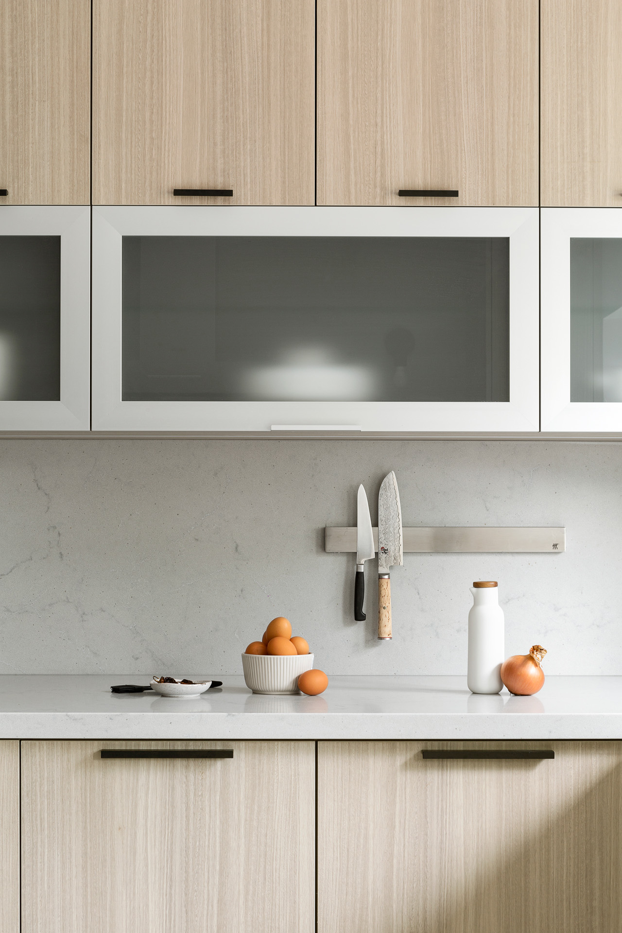 A close-up view of a modern kitchen featuring flat-panel light wood cabinetry accented with minimalist matte black edge pulls. A horizontal white-framed frosted glass cabinet is positioned above a light grey quartz countertop and matching seamless slab backsplash. A stainless steel magnetic knife rack is mounted to the wall holding two kitchen knives, while a bowl of fresh brown eggs, an onion, and a white bottle are styled on the counter below.