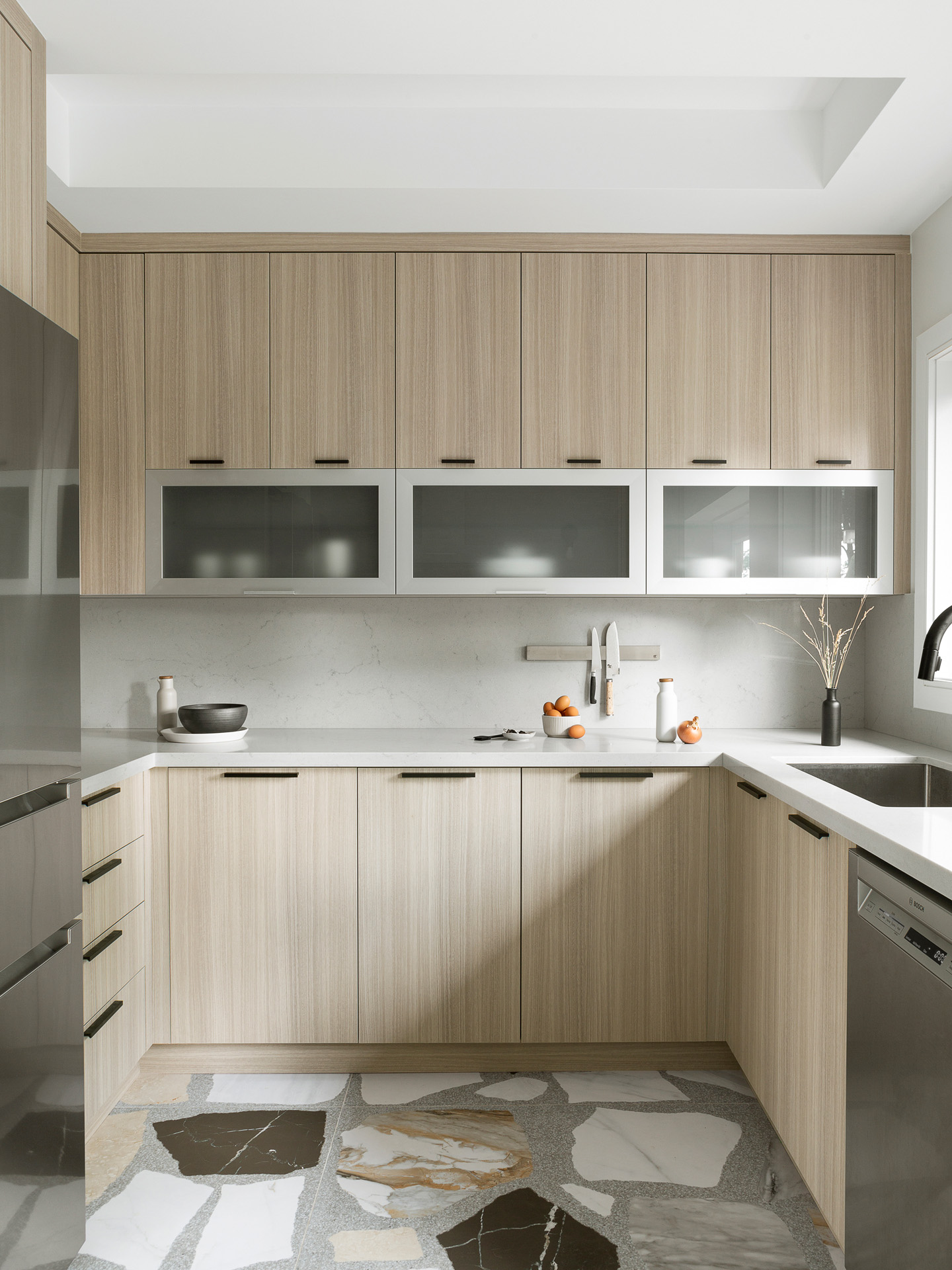 A bright, modern kitchen featuring flat-panel light wood cabinetry accented with minimalist matte black edge pulls. A unique row of horizontal frosted glass cabinets is positioned above a white quartz countertop and a matching seamless slab backsplash fitted with a magnetic knife strip. The space is grounded by a striking large-scale terrazzo floor featuring oversized fragments of white, black, and brown marble. Stainless steel appliances, including a refrigerator and dishwasher, complete the contemporary design.