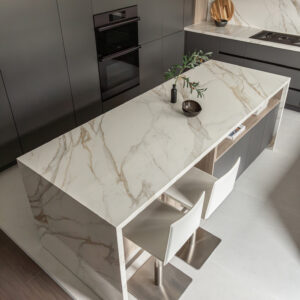 An elevated, angled view of a sleek modern kitchen highlighting a stunning white marble waterfall island with prominent gold and grey veining. Two modern white leather bar stools with square stainless steel bases sit at the island. The background features seamless floor-to-ceiling matte dark grey cabinetry with a built-in double wall oven, complemented by matching marble perimeter countertops and a full-height backsplash.