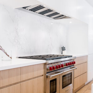 A close-up of a modern kitchen featuring a professional stainless steel gas range with distinctive red knobs, flanked by light wood flat-panel base cabinets. A seamless white marble-look quartz forms the countertop and full-height backsplash. Above the stove, a stainless steel range hood insert is discreetly integrated into sleek, minimalist white upper cabinetry.