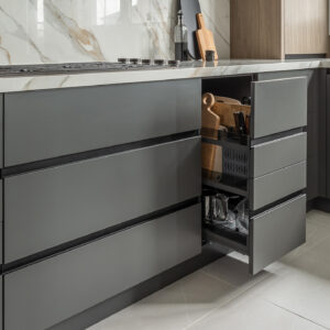 A close-up view of a modern kitchen's lower cabinetry featuring sleek, matte dark grey handleless drawers. One tall pull-out cabinet is open, revealing a highly organized interior with custom metal racks storing wooden cutting boards, utensils, and stainless steel pots. The cabinets sit beneath a white marble countertop with a built-in black gas cooktop and matching marble backsplash.
