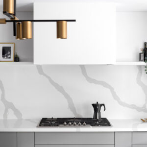 A modern kitchen featuring dark grey flat-panel base cabinets and a striking white quartz slab countertop and matching backsplash with bold, continuous grey veining. A sleek, minimalist white custom range hood is centered over a built-in gas cooktop. A single long white floating shelf spans the width of the backsplash, elegantly styled with framed artwork, oil bottles, and a trailing plant. A contemporary black and brass linear chandelier hangs in the foreground.