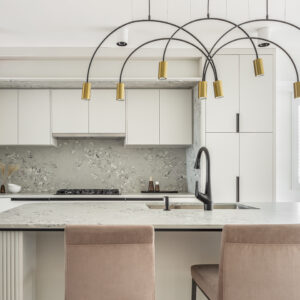 A bright, contemporary kitchen featuring a large central island with a speckled terrazzo-style countertop, white fluted edge detailing, and a matte black faucet, paired with dusty rose velvet bar stools. A striking modern chandelier with sweeping black arched arms and brass cylindrical shades hangs prominently above. The background showcases sleek off-white flat-panel cabinetry with black hardware, a matching continuous terrazzo backsplash, and built-in stainless steel wall ovens.