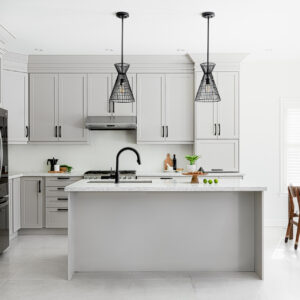 A bright, spacious modern kitchen featuring light grey shaker-style cabinets adorned with matte black hardware. A central island with a white speckled stone countertop and a matte black gooseneck faucet sits beneath two striking black wireframe pendant lights. The kitchen includes stainless steel appliances, light grey tile flooring, and seamlessly flows into a dining area on the right with a dark wood table and chairs beside sunlit windows.
