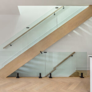 A sleek, modern staircase featuring a thick light oak stringer, matching handrails, and frameless glass balustrades secured with minimalist black metal hardware. Sunlight casts geometric shadows on the crisp white walls behind the stairs. The space is anchored by elegant light wood flooring laid in a chevron pattern, with a partial view of a white fluted cabinet in the right foreground.