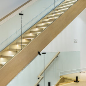 A close-up angled view of a modern interior staircase featuring light wood treads and a matching thick wooden stringer. The sleek, contemporary design is highlighted by frameless glass railings secured with minimalist black metal hardware and integrated warm LED strip lighting under each step. The illuminated stairs are set against clean white walls.