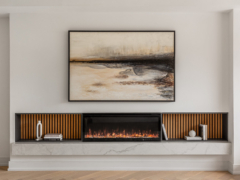 A modern living room focal point featuring a large framed abstract landscape painting in earth tones mounted above a sleek, built-in linear electric fireplace. The fireplace is symmetrically flanked by dark open shelving with vertical wood slat backings, minimally styled with books and white ceramic decor. The entire unit rests on a thick, floating white marble hearth over light wood flooring.